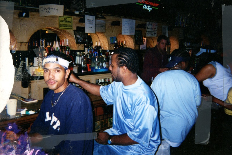Four patrons sitting at the bar in Soakie's. A masculine patron in the foreground looks straight into the camera, the others are in conversation.