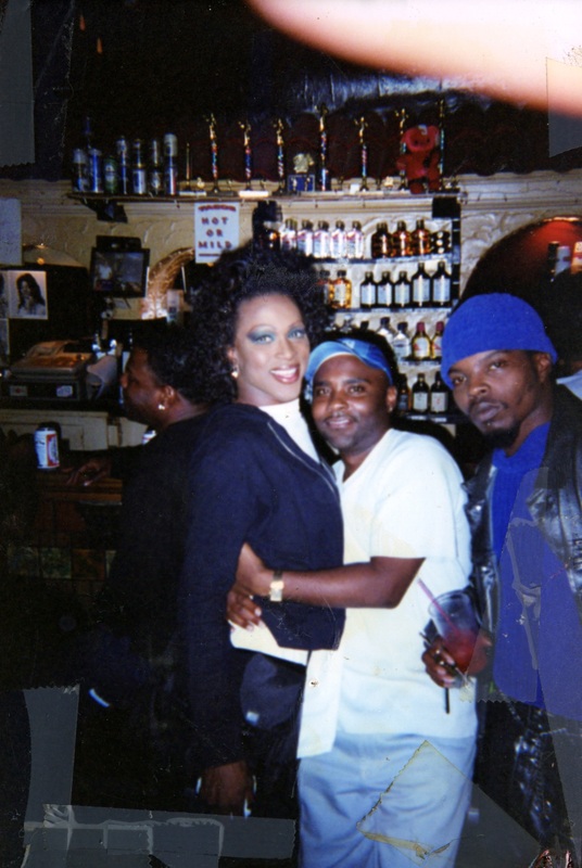 A feminine performer and two masculine patrons in front of the bar at Soakie's. One patron hugs the performer–both of them smile; the other looks straight on to the camera.