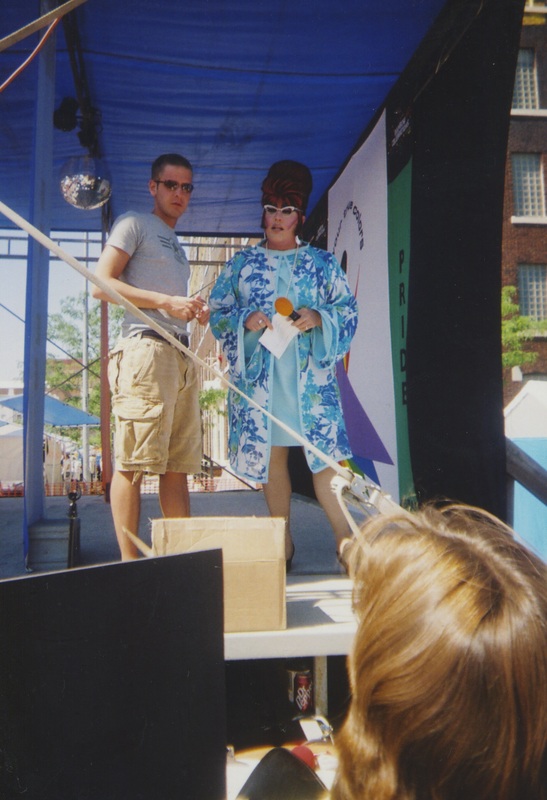 A white masculine volunteer and white drag queen, Flo, at Kansas City's PRIDE from an unknown year. Both look into the audience fixated on someone.