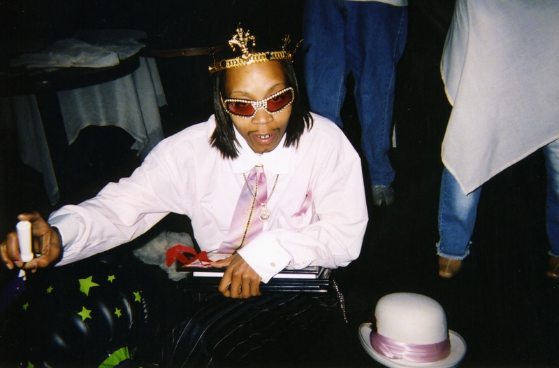 A masculine performer with a crown and bedazzled glasses looks into the lens at Soakie's.